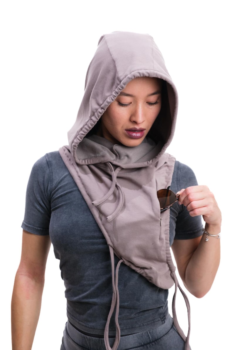 Op de afbeelding: Een kap sjaal in een gedempte mauve kleur, met een ingebouwde zak. De sjaal heeft trekkoorden en wordt over een grijs t-shirt gedragen. Zonnebrillen worden in de zak gestopt. Het ontwerp is casual en praktisch.