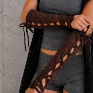 May include: A pair of dark brown arm warmers featuring a lace-up design with matching brown cord ties that run along the outer length of the sleeves. The fabric has a subtle textured pattern and extends from the upper forearm down to the wrist area.