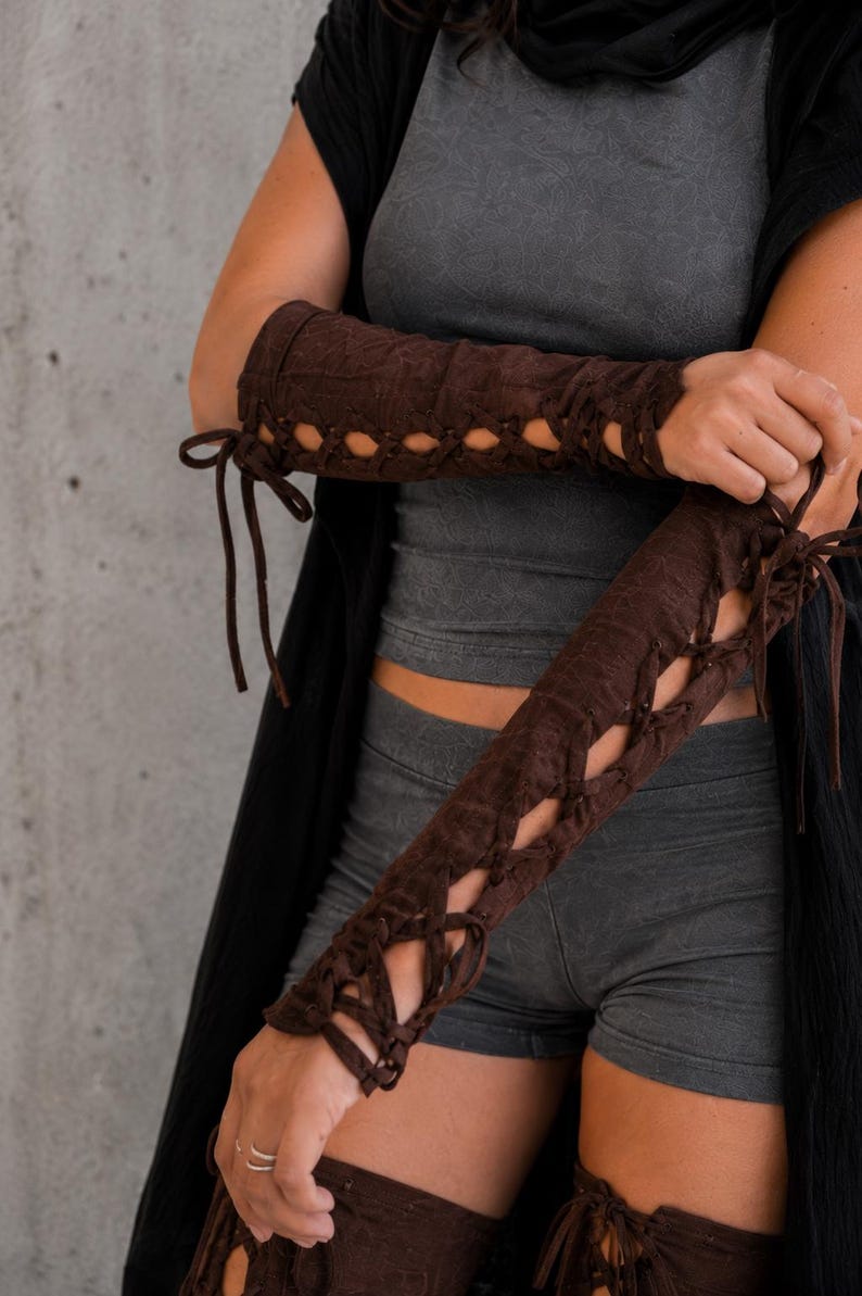 May include: A pair of dark brown arm warmers featuring a lace-up design with matching brown cord ties that run along the outer length of the sleeves. The fabric has a subtle textured pattern and extends from the upper forearm down to the wrist area.
