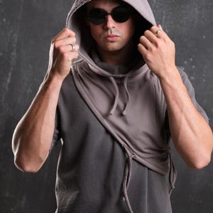 Op de afbeelding: Een grijze hoodie met een uniek asymmetrisch ontwerp. De capuchon is omhoog en het model draagt een donkere zonnebril. De sweatshirt heeft een trekkoorddetail en een gelaagde look, wat een moderne, stedelijke esthetiek cre&euml;ert.