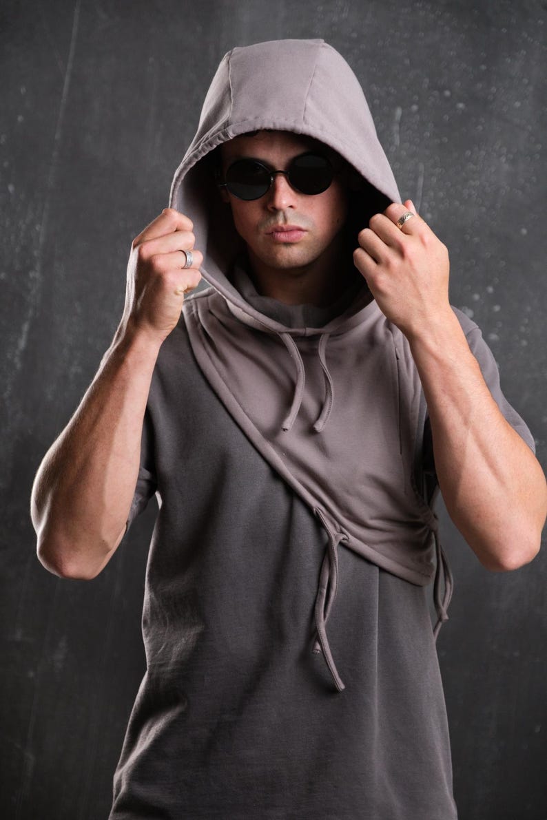 Op de afbeelding: Een grijze hoodie met een uniek asymmetrisch ontwerp. De capuchon is omhoog en het model draagt een donkere zonnebril. De sweatshirt heeft een trekkoorddetail en een gelaagde look, wat een moderne, stedelijke esthetiek cre&euml;ert.