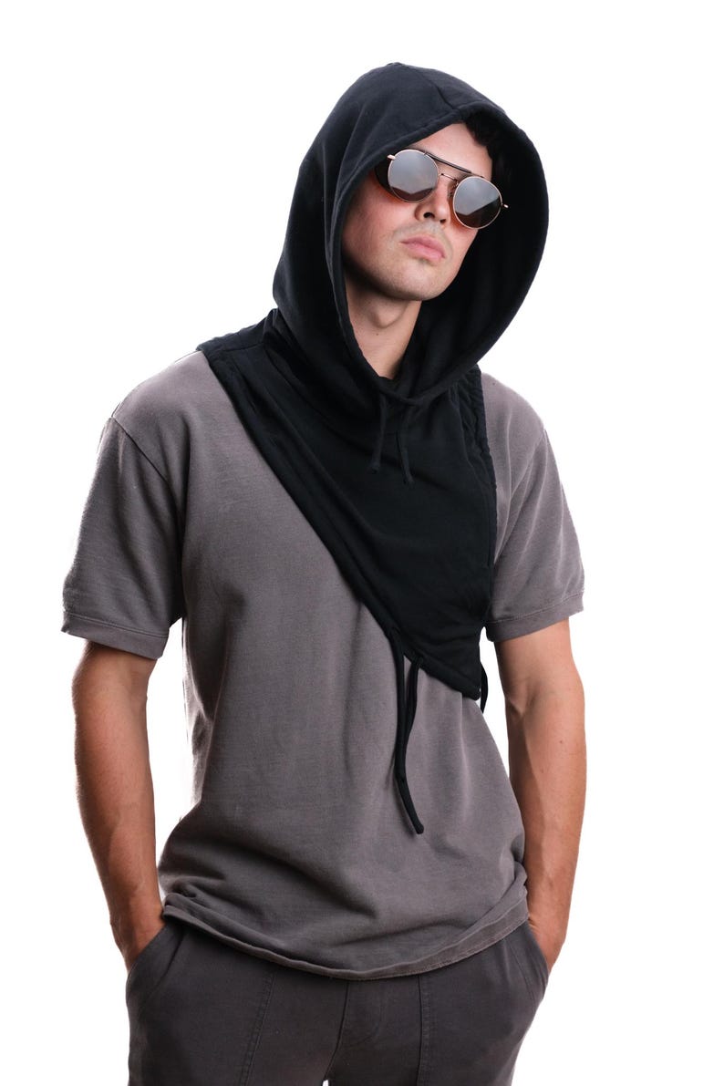 Op de afbeelding: Een zwarte sjaal met capuchon en trekkoord, gedragen over een grijs t-shirt. De sjaal is over de schouders gedrapeerd. De persoon draagt ​​ronde zonnebrillen.