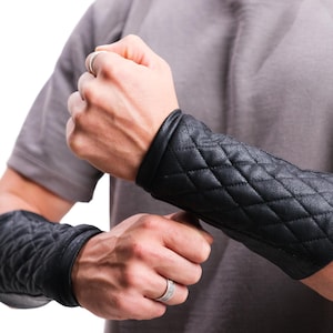 Könnte beinhalten: Schwarze Leder-Armstulpen mit einem gesteppten Rautenmuster. Die Stulpen werden an den Unterarmen getragen und haben einen glatten, abgerundeten Rand. Die Stulpen sind von tiefschwarzer Farbe.