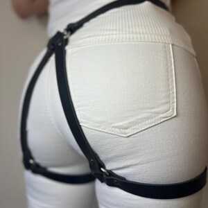 May include: A black leather-like harness accessory featuring a waist strap connected to two adjustable thigh straps by vertical bands. The harness is constructed with silver-toned metal rings and rivets, designed to be worn over white denim pants.