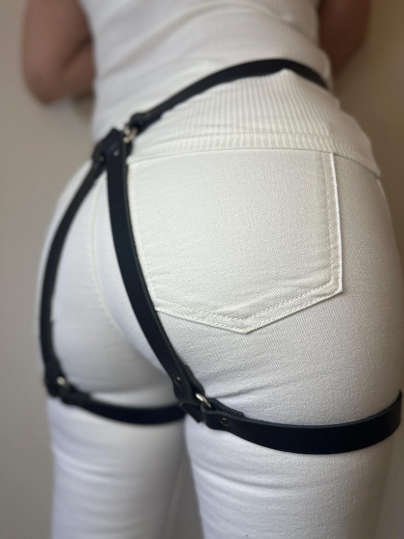 May include: A black leather-like harness accessory featuring a waist strap connected to two adjustable thigh straps by vertical bands. The harness is constructed with silver-toned metal rings and rivets, designed to be worn over white denim pants.
