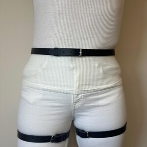 May include: A black leather belt worn around the waist and two matching black leather leg straps worn around the thighs. Each strap features a silver-toned metal buckle and multiple adjustment holes for a secure fit against the white clothing.