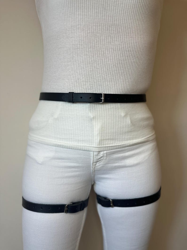 May include: A black leather belt worn around the waist and two matching black leather leg straps worn around the thighs. Each strap features a silver-toned metal buckle and multiple adjustment holes for a secure fit against the white clothing.