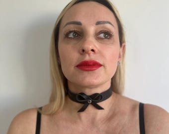 May include: Black choker necklace with a bow detail. The choker is made of a smooth, dark material and features a small bow centered at the front. A small, silver-colored stud is in the center of the bow.