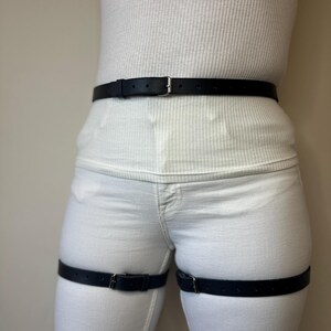 May include: A black leather belt worn around the waist and two matching black leather straps worn around each thigh. Each strap features a silver-toned metal buckle and multiple adjustment holes, providing a secure fit over white clothing.