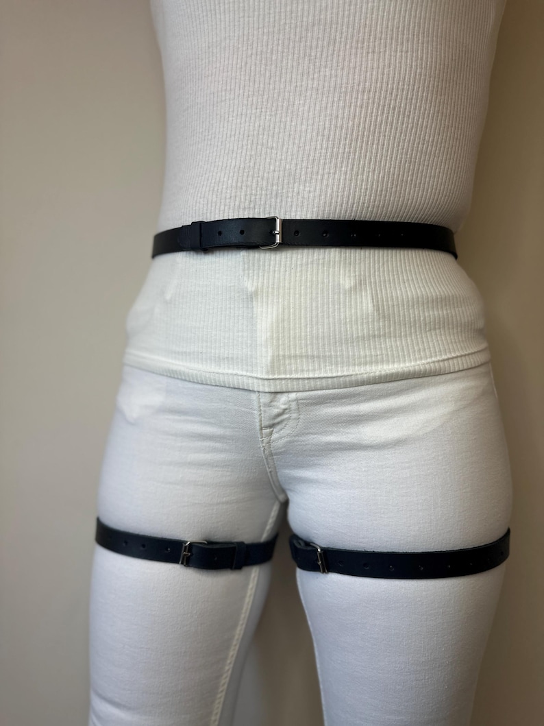 May include: A black leather belt worn around the waist and two matching black leather straps worn around each thigh. Each strap features a silver-toned metal buckle and multiple adjustment holes, providing a secure fit over white clothing.