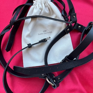 May include: A black leather harness featuring adjustable straps with silver-toned metal hardware and buckles. The harness is displayed alongside a cream-colored fabric drawstring pouch printed with the text LSD Leather Star Design.