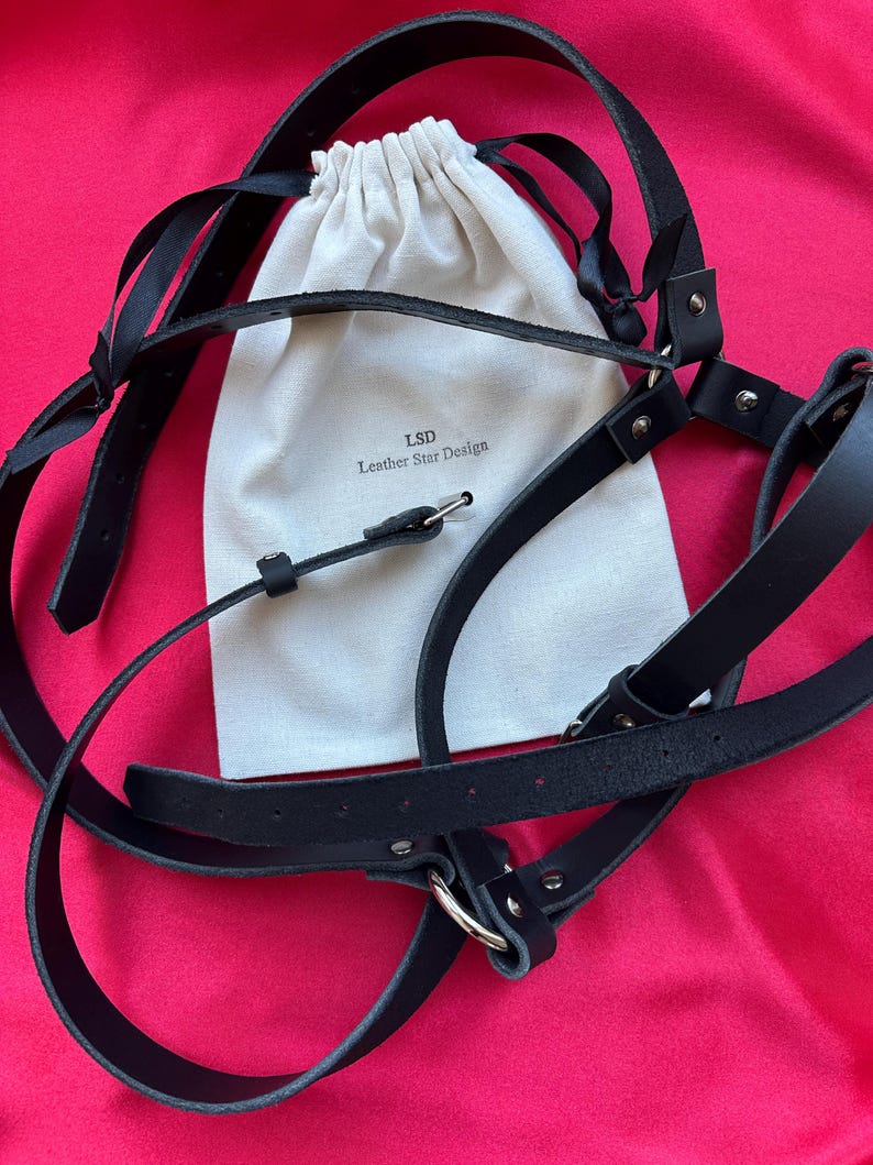 May include: A black leather harness featuring adjustable straps with silver-toned metal hardware and buckles. The harness is displayed alongside a cream-colored fabric drawstring pouch printed with the text LSD Leather Star Design.