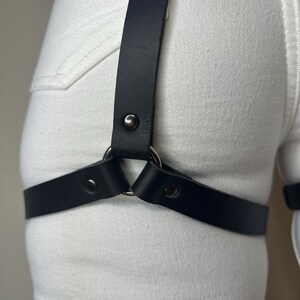 May include: A black leather leg harness featuring a central silver-toned metal ring that connects a vertical strap to a horizontal thigh strap. The straps are secured with matching silver-toned metal rivets, worn over white denim fabric.