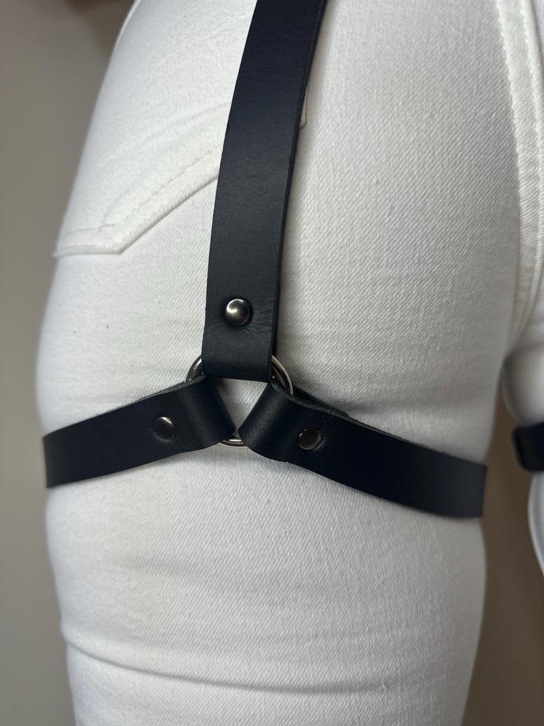May include: A black leather leg harness featuring a central silver-toned metal ring that connects a vertical strap to a horizontal thigh strap. The straps are secured with matching silver-toned metal rivets, worn over white denim fabric.