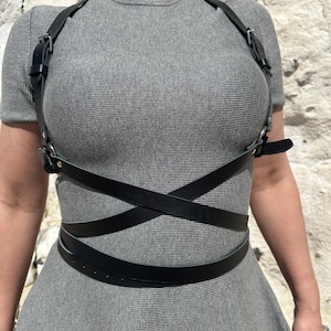 May include: Black leather body harness with adjustable straps and buckles. The harness has a crisscross design across the torso and a belt around the waist. The harness is worn over a grey dress.