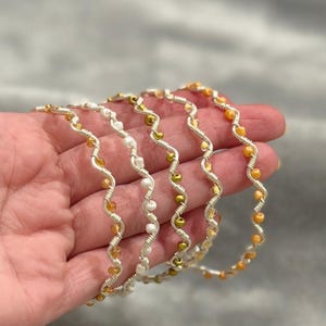 May include: A hand holding five silver-toned, wavy bangles. Each bangle is adorned with small, colorful beads in shades of gold, white, and orange. The bangles have a delicate, handcrafted appearance, suggesting they are jewelry.