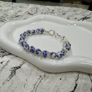 May include: A delicate bracelet featuring white beads with blue floral patterns, strung together with silver-colored wire. The bracelet is displayed on a white, organically shaped ceramic dish, set against a patterned fabric background.