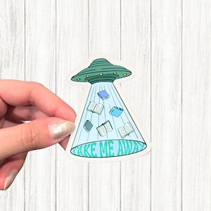 以下が含まれることがあります： 緑と青緑色のUFOが本を放出しているステッカー。ビームには開いた本と閉じた本があります。テキスト「TAKE ME AWAY」はビームの下部に書かれています。
