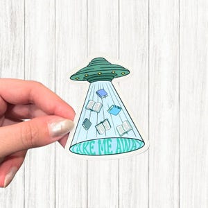 以下が含まれることがあります： 緑とターコイズ色のUFOが本を照らしているステッカー。UFOは、開いた本と閉じた本が入った水色の光線を持っています。「TAKE ME AWAY」というテキストがステッカーの下部に緑とターコイズ色で書かれています。