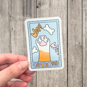 May include: A blue and white sticker with a cartoon paw print and the text "Woof!" and "Woof!" on a light blue background. The text "The Toe Beans" is at the bottom of the sticker.