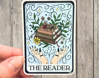 May include: A blue and white sticker with a whimsical design featuring two hands holding a stack of books, flowers, and stars. The text "THE READER" is printed at the bottom.