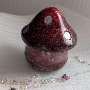 May include: A red and white mushroom-shaped trinket box with a glittery finish. The box is made of resin and has a white interior. The box is closed and sitting on a white surface.