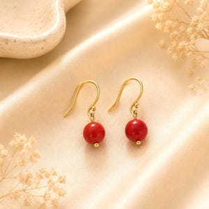 May include: A pair of gold-toned hook earrings featuring round, red, marbled beads. Each earring consists of a curved gold wire hook attached to a small gold jump ring, which holds a single spherical red bead with subtle dark veining throughout.