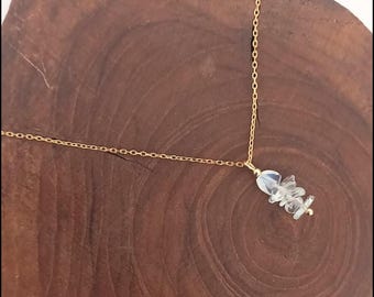 May include: A delicate gold-colored chain necklace featuring a cluster of clear, irregular-shaped stones. The necklace is displayed on a dark brown wooden surface, creating a contrast that highlights the jewelry's details.