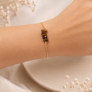 Può includere: Un delicato bracciale a catena color oro con un gruppo centrale di quattro piccole pietre preziose occhio di tigre levigate, nei toni del marrone e del giallo dorato. Il bracciale è formato da una sottile catena metallica che avvolge il polso.