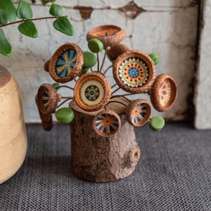 May include: A decorative arrangement featuring multiple acorn caps mounted on thin wire stems, each painted with intricate geometric patterns in orange, blue, and black. The stems are inserted into a small, cylindrical wooden log base.