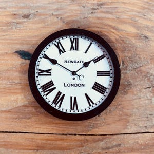 Puede incluir: Reloj redondo con marco negro y esfera blanca con números romanos y manecillas negras. La esfera del reloj dice "NEWGATE LONDON". El reloj está sobre un fondo de madera.