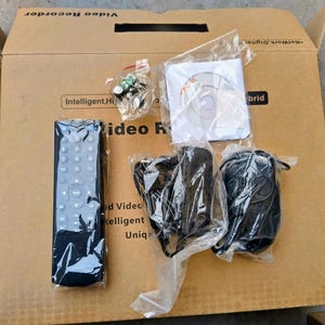 Puede incluir: Una caja de cartón que contiene una grabadora de vídeo y sus accesorios. Los accesorios incluyen un mando a distancia, un CD, cables y pequeños componentes electrónicos, todos en embalaje de plástico. La caja tiene texto impreso.