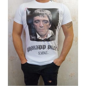 Könnte beinhalten: Ein weißes kurzärmeliges T-Shirt mit einem großen rechteckigen Grafikdruck eines Männergesichts auf der Brust. Unter dem Bild steht der Text PHILIPP PLEIN in einer gotischen Schrift, gefolgt von dem Wort SCARFACE in einer serifenlosen Schrift.