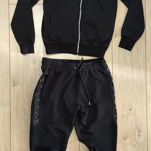Può includere: Completo tuta nero con giacca con zip e pantaloni abbinati. La giacca presenta un logo sul petto e dettagli a fantasia sulle maniche. I pantaloni hanno un girovita con coulisse e cerniere alle caviglie. Il completo è realizzato in un tessuto scuro e liscio.