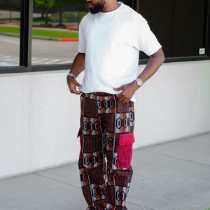 May include: A plain white short-sleeved t-shirt paired with loose-fitting cargo pants featuring a geometric pattern in shades of brown, black, and grey, accented by solid red side pockets and red-trimmed black and white athletic sneakers.
