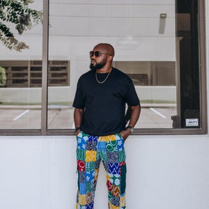 May include: A black short-sleeved t-shirt paired with vibrant, multi-colored patchwork trousers featuring various geometric and floral patterns in shades of blue, yellow, red, green, and white, worn with light-colored sneakers and a gold chain necklace.