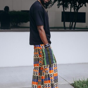 May include: A black short-sleeved t-shirt paired with wide-leg trousers featuring a vibrant, multi-colored geometric pattern in orange, blue, white, and black, accented by a vertical green and black striped patch pocket on the side.