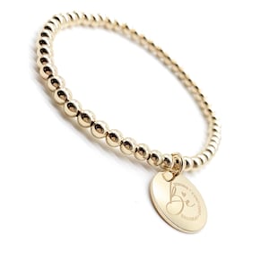 Puede incluir: Pulsera dorada de cuentas con un dije redondo. El dije está grabado con la palabra "Be" y las palabras "Strong, Confident, Positive". La pulsera está sobre un fondo blanco.
