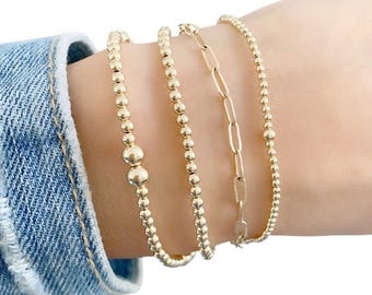 May include: A set of four gold-toned bracelets worn together. Three bracelets feature small round beads, with one including two larger central beads. The fourth bracelet consists of elongated oval links. The bracelets are worn on a wrist.