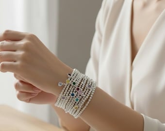 May include: Silver beaded bracelets with colorful gemstone accents. The bracelets are stacked on a wrist, showcasing a variety of colors including green, red, and purple. A single charm with a clear stone hangs from one bracelet.
