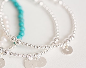 May include: Close-up of several beaded bracelets. The bracelets are silver and have small, round beads. Some bracelets have small, round, silver charms with letters on them. One bracelet is turquoise. The bracelets are on a white surface.