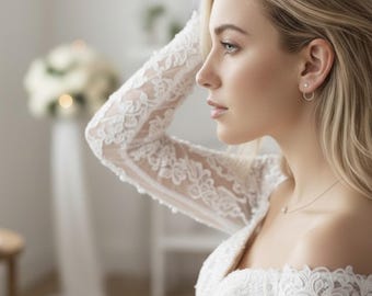 May include: A bride in a white lace wedding dress with off-the-shoulder sleeves. The dress features intricate floral lace detailing. The bride is wearing small hoop earrings and a delicate necklace. Her blonde hair is styled down. A bouquet of white flowers is in the background.