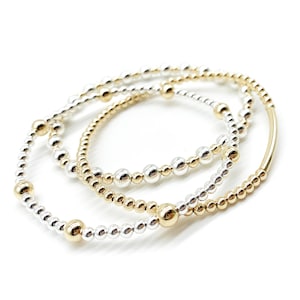 May include: Three beaded bracelets, one silver and two gold, stacked on top of each other.