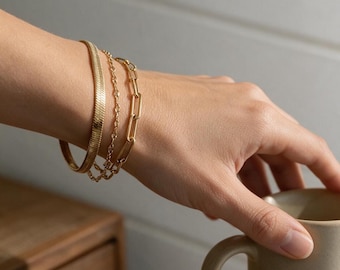 May include: A set of three gold-toned bracelets worn together. The stack includes a flat herringbone chain bracelet, a delicate twisted rope chain bracelet, and a larger rectangular paperclip link chain bracelet, all featuring a polished finish.