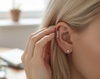 May include: A close-up of a person's ear adorned with a silver ear cuff featuring three rows of small, sparkling stones. A single, small stud earring is also visible. The person's blonde hair is partially visible, and their hand gently touches the ear.