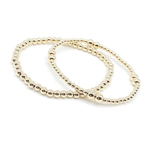 Op de afbeelding: Twee gouden kralen armbanden, een iets groter dan de andere, liggen op elkaar op een witte achtergrond.