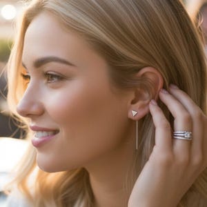 May include: A pair of silver earrings featuring a small triangle stud and a delicate chain that dangles down. The earrings are worn by a person. A silver ring with multiple bands is also visible.
