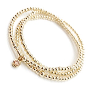 May include: A gold beaded bracelet with a single pink gemstone charm. The bracelet is made of three strands of small gold beads.