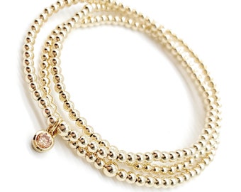 May include: A multi-strand bracelet composed of three rows of small, polished gold-toned spherical beads. A single, circular charm featuring a light peach-colored gemstone set in a gold-toned bezel hangs from one of the beaded strands.