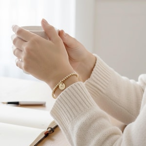 Op de afbeelding: Een gouden kralenarmband met een kleine ronde bedel met het nummer 40. De armband wordt om een pols gedragen en de persoon houdt een witte mok vast. De armband is een eenvoudig, elegant sieraad.